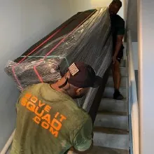 A Move It Squad mover in a company t-shirt carrying a wrapped sofa up the stairs